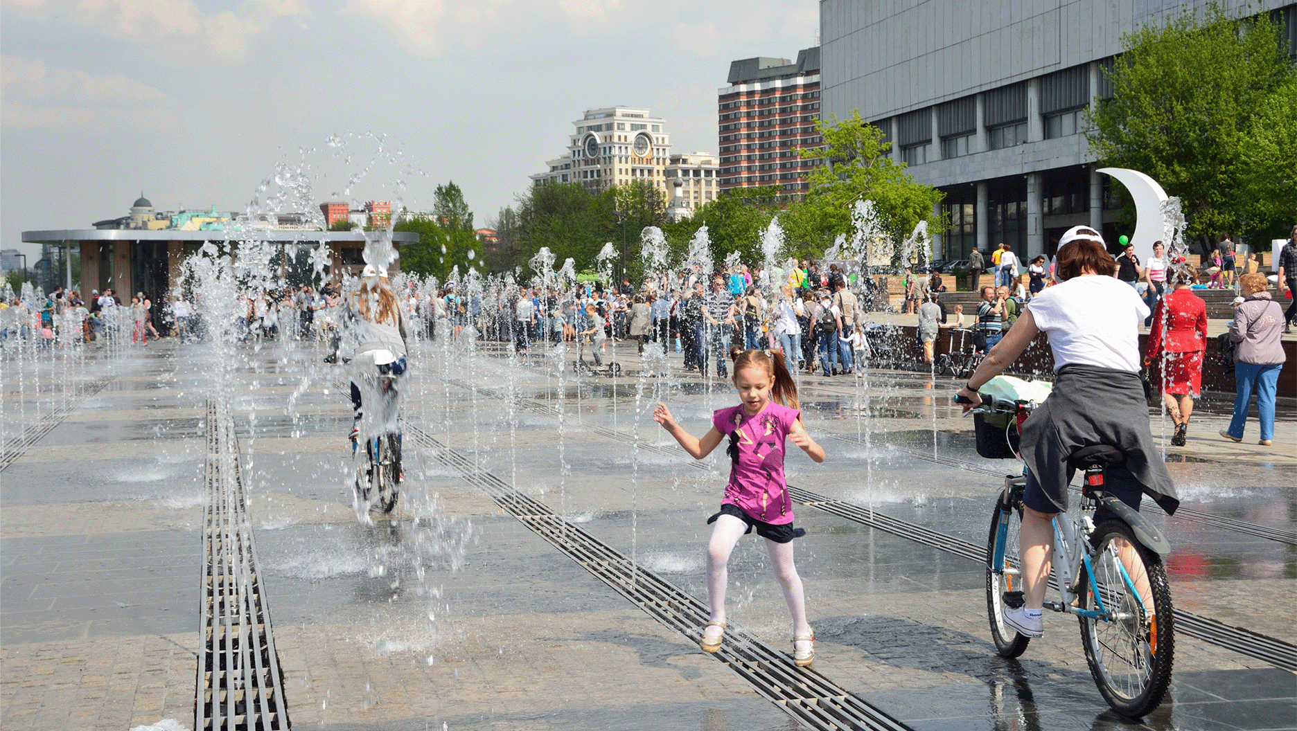 En été, des passants se rafraîchissent sous des jets d’eau d’une grande place publique.