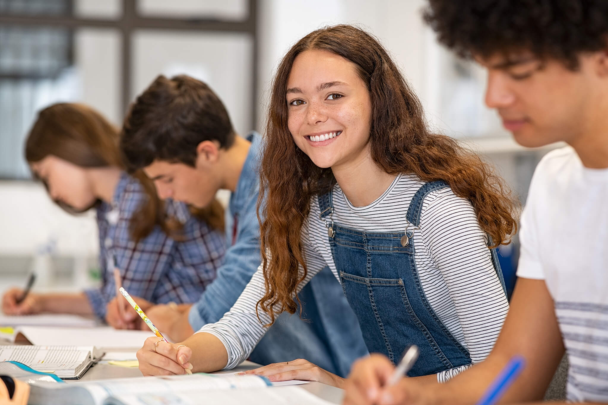 La photo montre une élève dans une classe. L’adolescente a un crayon à la main et est occupée à écrire dans son cahier, comme les trois autres élèves que l’on aperçoit autour d’elle.