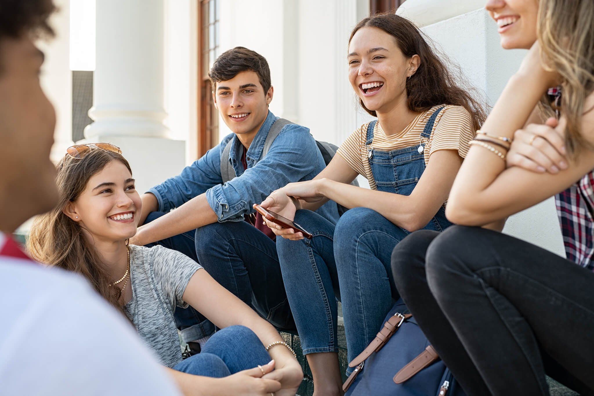 La photo montre la même adolescente que dans la photo précédente assise sur des marches et installée au milieu d’un groupe d’amis filles et garçons. Elle rit alors qu’elle discute avec ses amis qui rient aussi.