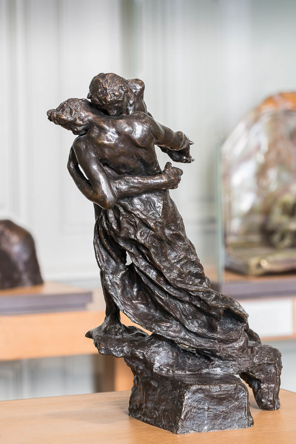 Sculpture de bronze d’environ 40 centimètres de hauteur. Un homme et une femme s’enlacent en dansant joue contre joue. Ils sont vêtus d’un tissu drapé qui couvre le bas de leur corps.